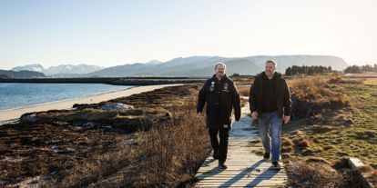 To menn går på sti ved sjøen. Det er fjell i bakgrunnen. 