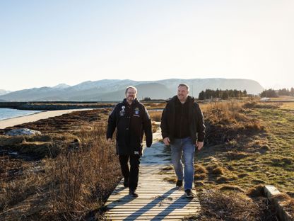 To menn går på sti ved sjøen. Det er fjell i bakgrunnen. 