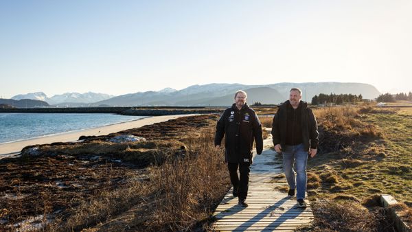 To menn går på sti ved sjøen. Det er fjell i bakgrunnen. 