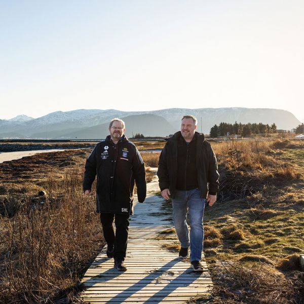 To menn går på sti ved sjøen. Det er fjell i bakgrunnen. 