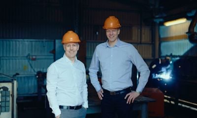 To forretningsmenn med oransje vernehjelmer i industrilokale. De representerer næringslivet og samarbeid for vekst og innovasjon i industribransjen.