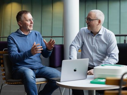 To menn i samtale om Aidency 113KIT – en innovasjon innen akuttmedisinsk støtte. En bærbar medisinsk koffert og en MacBook er på bordet, mens de diskuterer fremtidens akutthjelp.