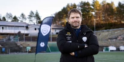 Mann står på en fotballbane, har armene i kryss og smiler