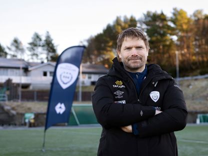 Mann står på en fotballbane, har armene i kryss og smiler