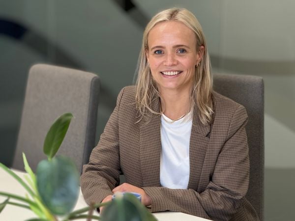 En dame med blondt hår og dressjakke sitter ved et bord og smiler til kamera