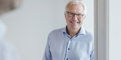 Mann i blå skjorte lener seg mot vinduskarm, ser i kamera og smiler. 