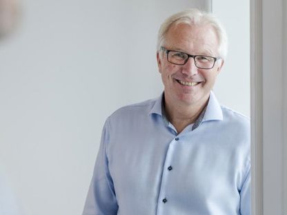 Mann i blå skjorte lener seg mot vinduskarm, ser i kamera og smiler. 