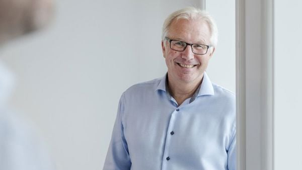 Mann i blå skjorte lener seg mot vinduskarm, ser i kamera og smiler. 