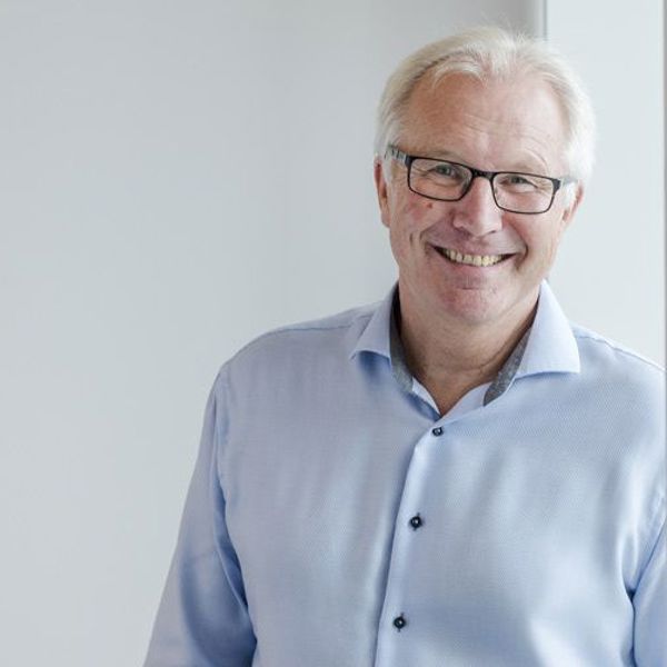 Mann i blå skjorte lener seg mot vinduskarm, ser i kamera og smiler. 
