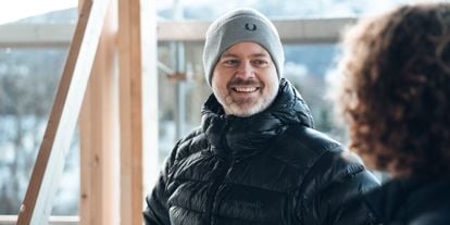 En mann med grå lue og svart jakke smiler til en person i et hus under bygging