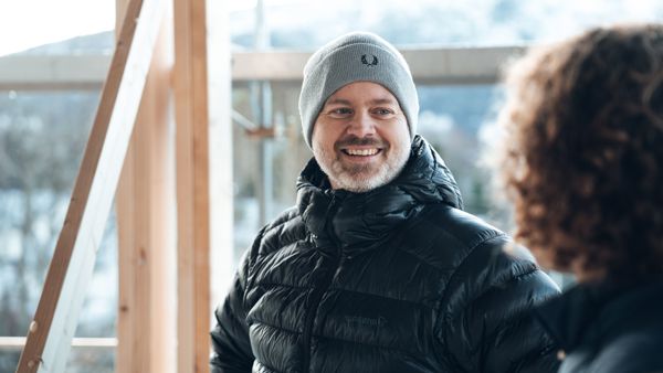 En mann med grå lue og svart jakke smiler til en person i et hus under bygging