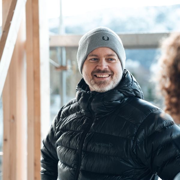 En mann med grå lue og svart jakke smiler til en person i et hus under bygging