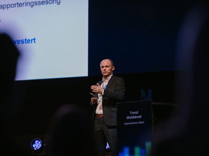 Mann med dress, står på en scene og presenterer. 