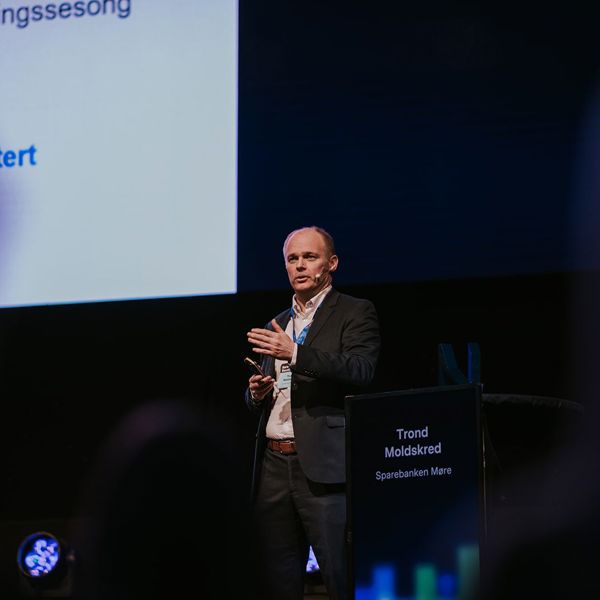 Mann med dress, står på en scene og presenterer. 