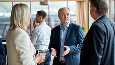 Flere mennesker i minglesituasjon. Mann i dress står i midten og er i fokus i bildet. 