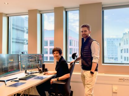 En kvinne sitter på kontorstol med skjermer med grafer framfor seg. En mann står bak stolen. Begge ser i kamera. Byutsikt i vinduene bak.