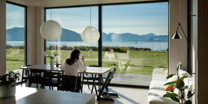 Kvinne sitter ved kjøkkenbord, ser ut av vindu mot havet.