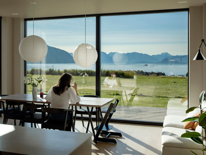 Kvinne sitter ved kjøkkenbord, ser ut av vindu mot havet.