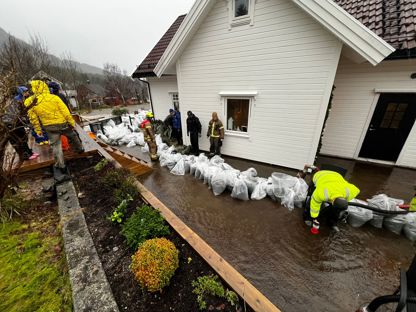 Bildet viser flere mennesker i en hage som er oversvømmet med vann. Langs huskanten ligger metervis med sandsekker stablet opp. 