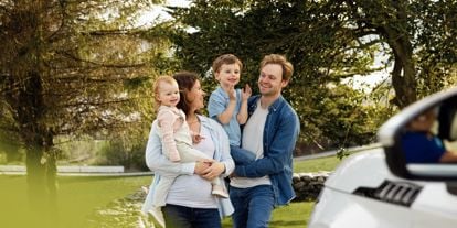 Familie ute, fremfor en bil. Mor, med datter på armen. Far med sønn på armen. 