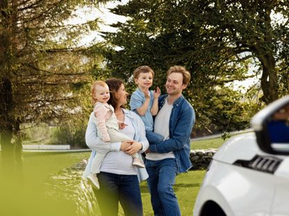 Familie ute, fremfor en bil. Mor, med datter på armen. Far med sønn på armen. 
