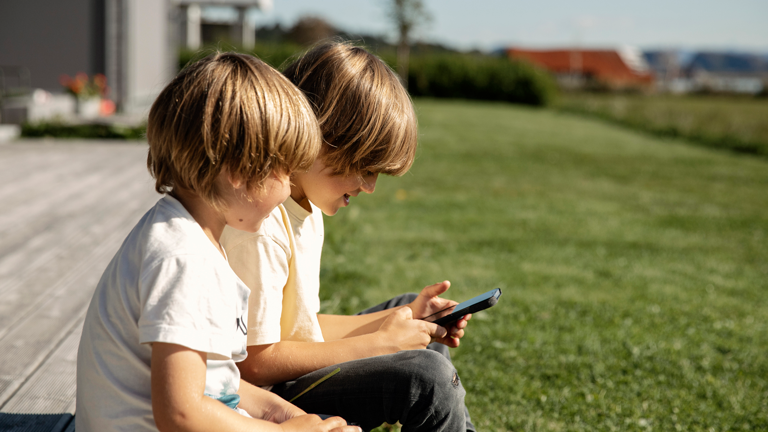 To gutter sitter på terrasse, den ene gutten holder en mobiltelefon begge ser på.