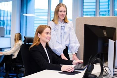 En kvinne kledd i sort dressjakke sitter ved kontorpult. Kvinne i lys bluse står ved siden av. De ser på skjermen. 