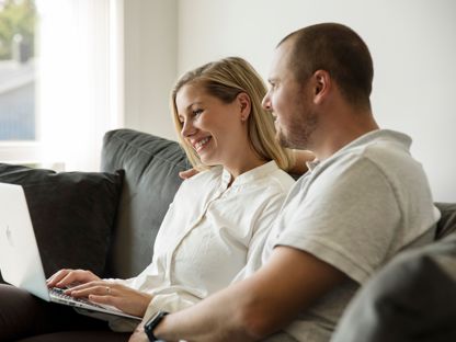 Mann og dame i lyse klær sitter i en mørk sofa med laptop på fanget. De ser på skjermen sammen. 