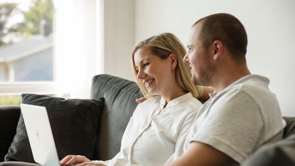 Mann og dame i lyse klær sitter i en mørk sofa med laptop på fanget. De ser på skjermen sammen. 