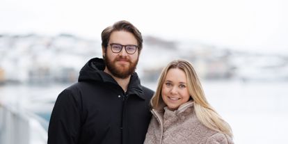 Ungt par som smiler til kamera, vinterkledd bylandskap i bakgrunnen 