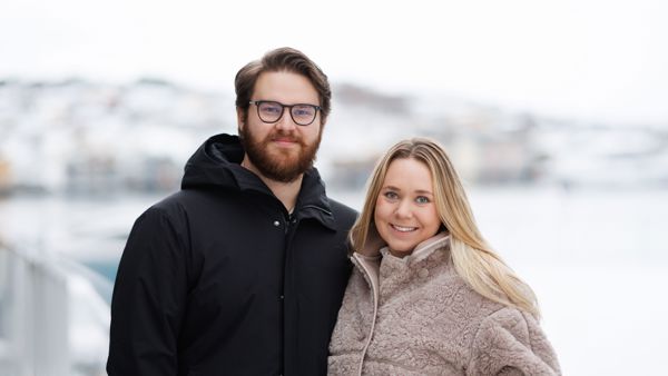 Ungt par som smiler til kamera, vinterkledd bylandskap i bakgrunnen 