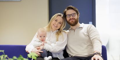Ung par med baby på fanget, sitter på blå sofa i et kontorlokale. 