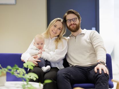 Ung par med baby på fanget, sitter på blå sofa i et kontorlokale. 
