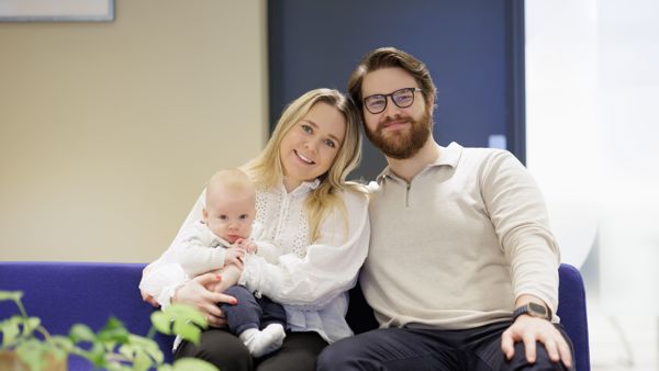 Ung par med baby på fanget, sitter på blå sofa i et kontorlokale. 