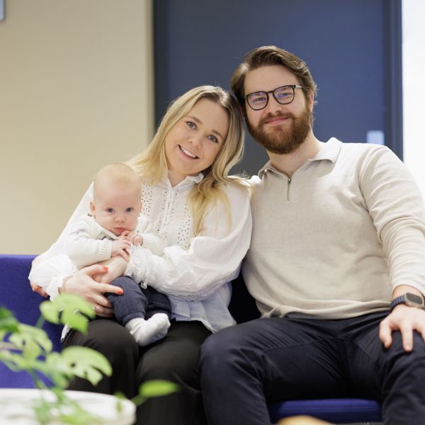 Ung par med baby på fanget, sitter på blå sofa i et kontorlokale. 
