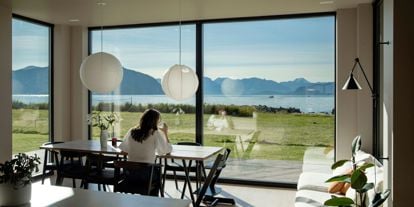 Kvinne sitter ved bord, drikker kaffe og ser ut av stort vindu med utsikt mot havet.