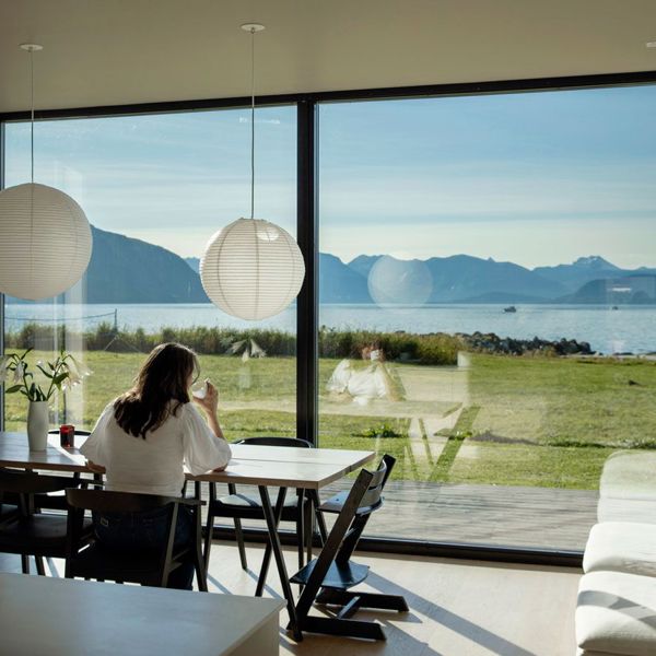Kvinne sitter ved bord, drikker kaffe og ser ut av stort vindu med utsikt mot havet.