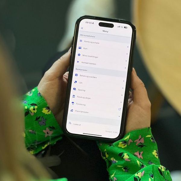 Person holder en smarttelefon med nettbankmeny på skjermen, mens de sitter ved et bord.