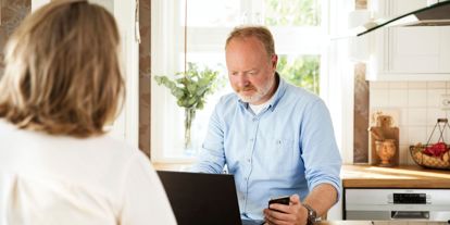 Bilde fra et kjøkken. En voksen mann sitter ved en kjøkkenøy, ser på en laptop og holder en mobil i hånden. Kredittkort ligger på benken.