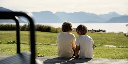 To barn sitter med ryggen til på en terrasse. De ser mot hav og fjell i horisonten. 