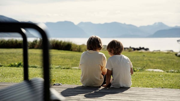 To barn sitter med ryggen til på en terrasse. De ser mot hav og fjell i horisonten. 