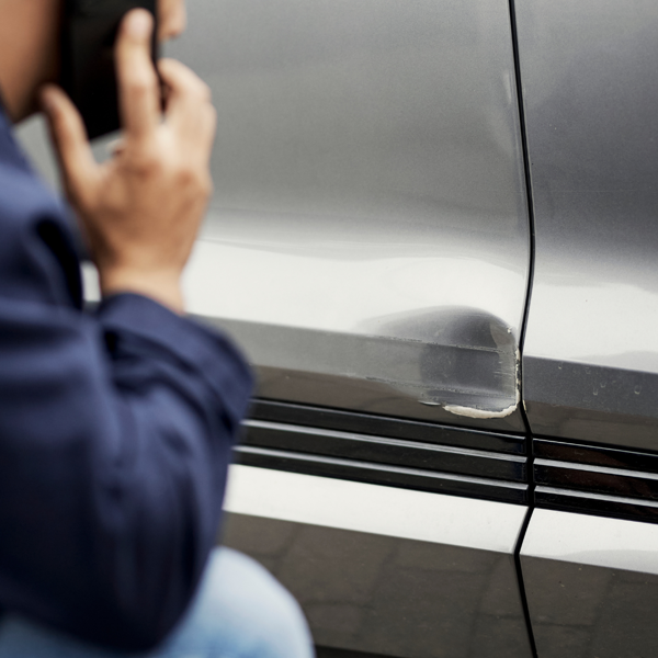 Bildet viser siden på en sølvfarget bil som har fått en bulk. Foran bilen sitter en person på huk.