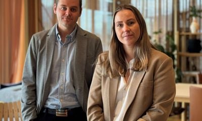 Kvinne sitter på bord, ser inn i kamera med betryggende blikk. Mann står litt bak kvinnen, ser også inn i kamera med betryggende blikk. 