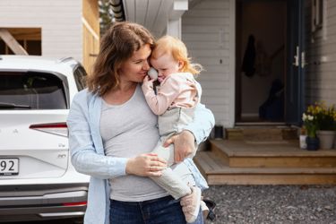 Gravid dame som holder en liten jente. Hus og bil i bakgrunnen. 