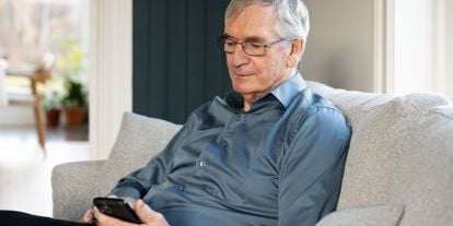 Eldre mann med blå skjorte, sitter i sofa og ser i mobiltelefonen. 