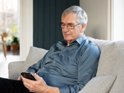 Eldre mann med blå skjorte, sitter i sofa og ser i mobiltelefonen. 