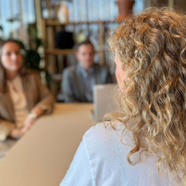 Bildet viser tre personer rundt et bord. I front er det en kvinne med blondt hår som sitter med ryggen mot kamera. På andre siden av bordet sitter en mann og en kvinne. De har blikket på kvinnen i front. 