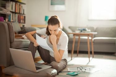 Kvinne sitter på stuegulvet, ser bekymret inn i skjermen på en laptop hun har fremfor seg. Holder et papir i hånden.