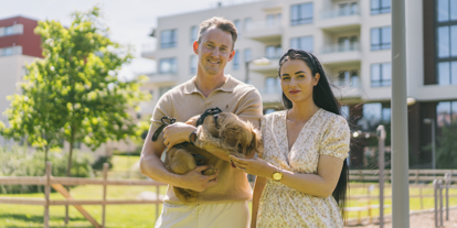 Dame og mann som står med en hund i hendene utenfor et boligområde i byen. Sol og sommer. 