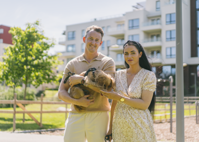 Et par står ute i solen og holder en liten hund foran moderne leilighetsbygg.
